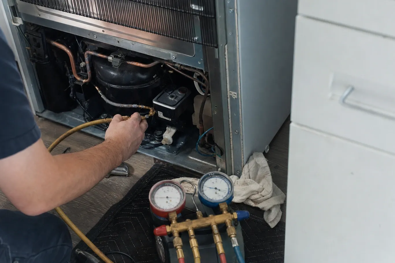 A hand on a yellow charging hose made up at the compressor of a built-in refrigerator with its lower panel off, the manifold set down on a mat on the floor